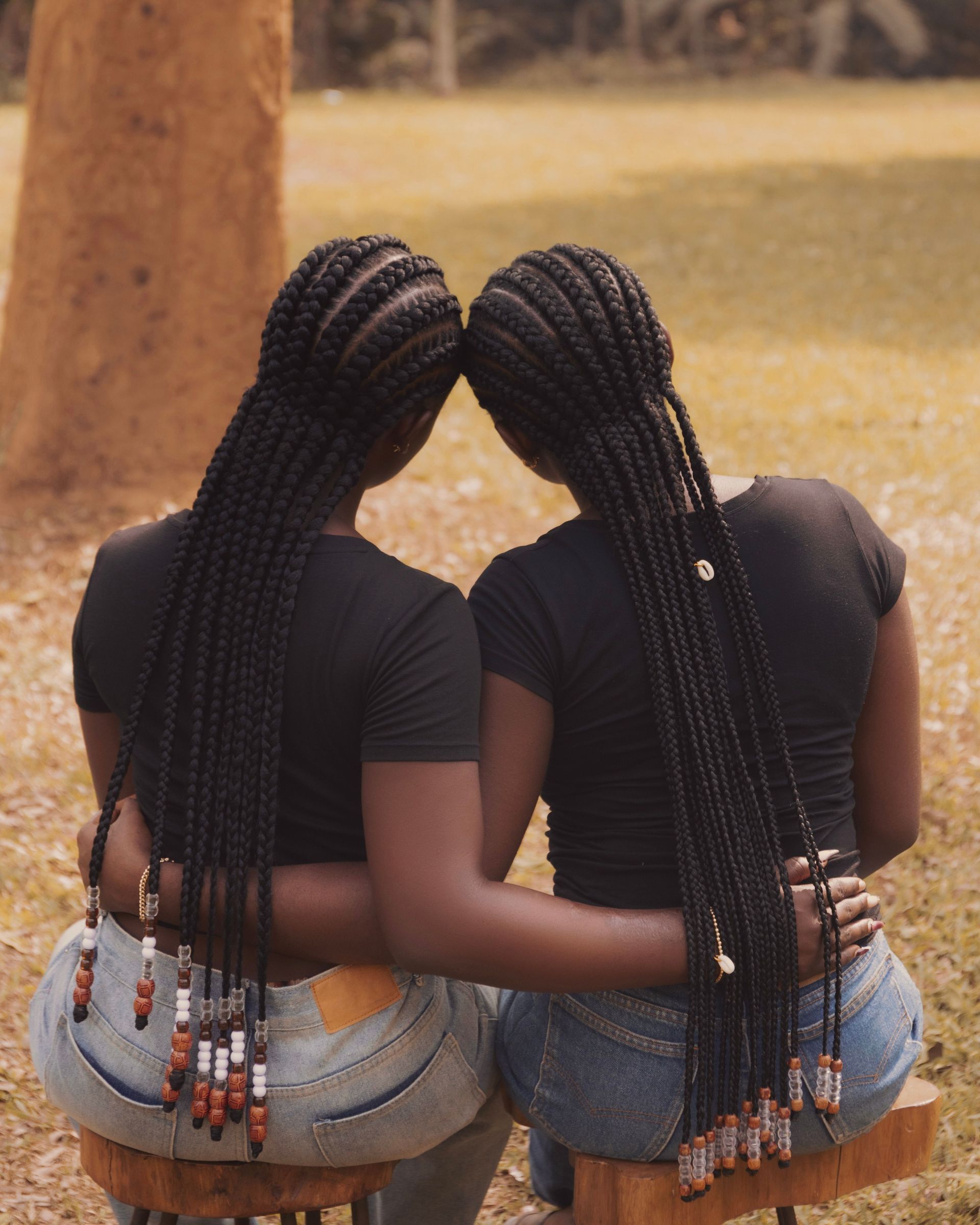 Two young women with long braided hair sitting outdoors, embracing and enjoying a sunny day.